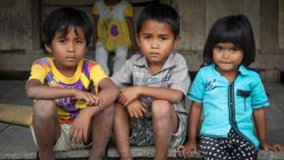 Three children sitting together outside a wooden home