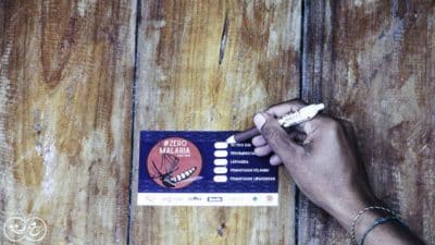 A hand writing on a Zero Malaria sticker attached to a wooden wall