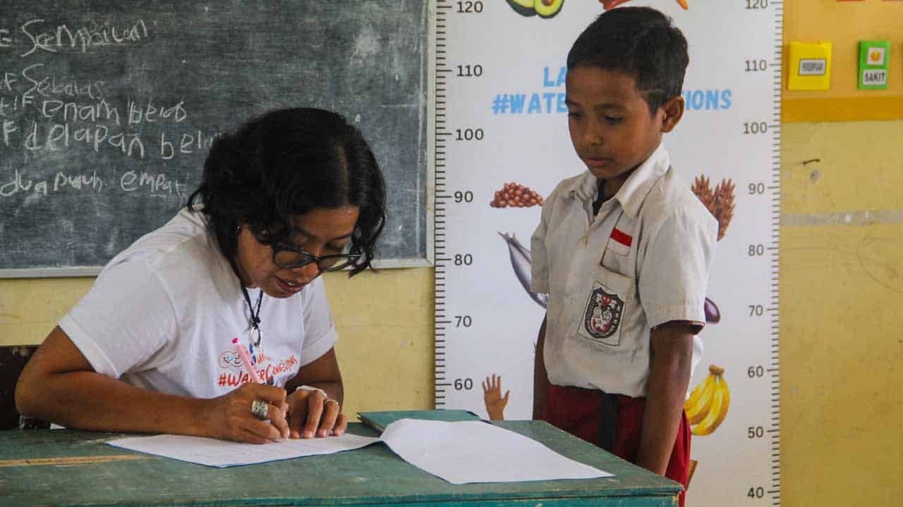 A health worker recording a schoolboy's measurements beside a height chart