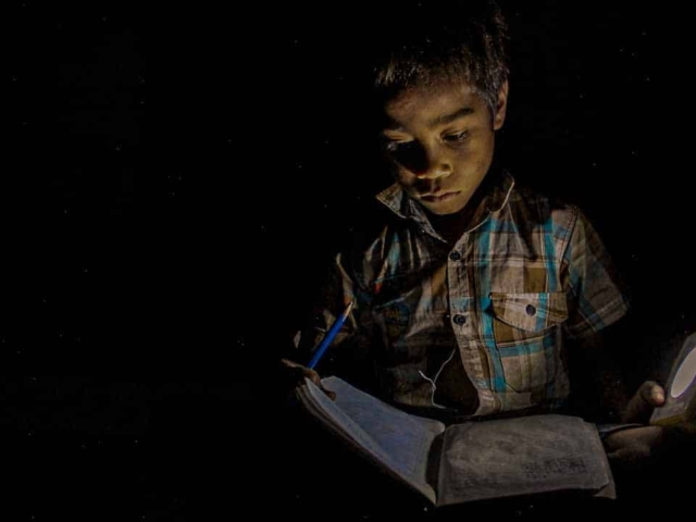 Children in East Sumba studying by the light of a SolarBuddy lamp, seated on the floor of their wooden home, deeply focused on their homework.