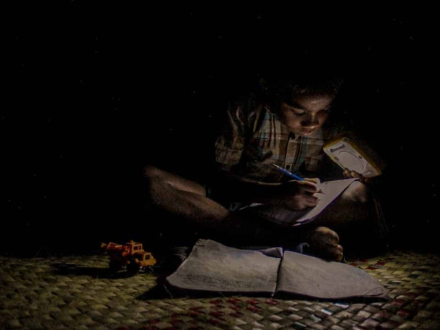 Children in East Sumba studying by the light of a SolarBuddy lamp, seated on the floor of their wooden home, deeply focused on their homework.