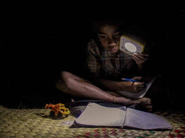 Children in East Sumba studying by the light of a SolarBuddy lamp, seated on the floor of their wooden home, deeply focused on their homework.