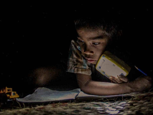 Children in East Sumba studying by the light of a SolarBuddy lamp, seated on the floor of their wooden home, deeply focused on their homework.