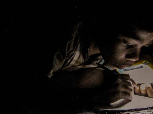 Children in East Sumba studying by the light of a SolarBuddy lamp, seated on the floor of their wooden home, deeply focused on their homework.