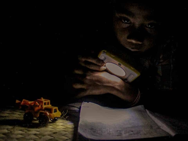 Children in East Sumba studying by the light of a SolarBuddy lamp, seated on the floor of their wooden home, deeply focused on their homework.