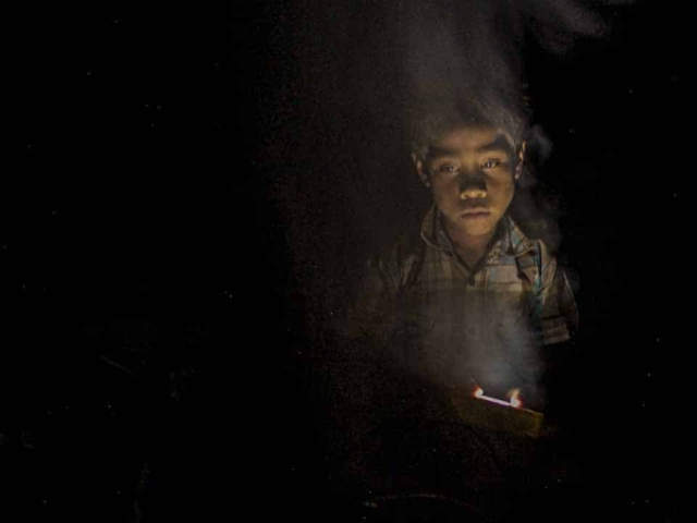 Children in East Sumba studying by the light of a SolarBuddy lamp, seated on the floor of their wooden home, deeply focused on their homework.