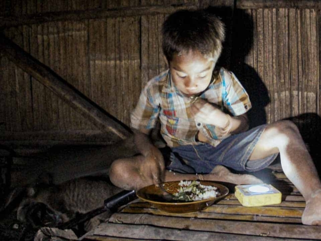 Children in East Sumba studying by the light of a SolarBuddy lamp, seated on the floor of their wooden home, deeply focused on their homework.