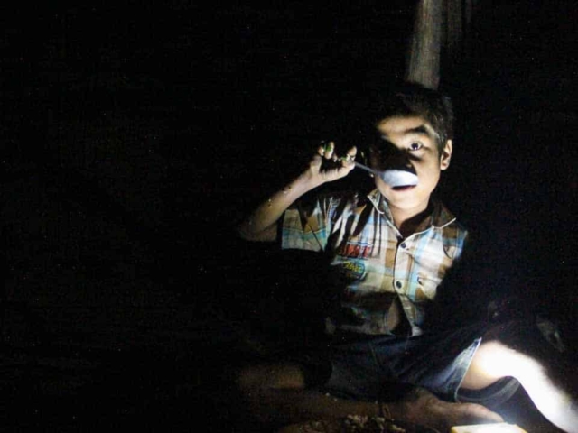 Children in East Sumba studying by the light of a SolarBuddy lamp, seated on the floor of their wooden home, deeply focused on their homework.