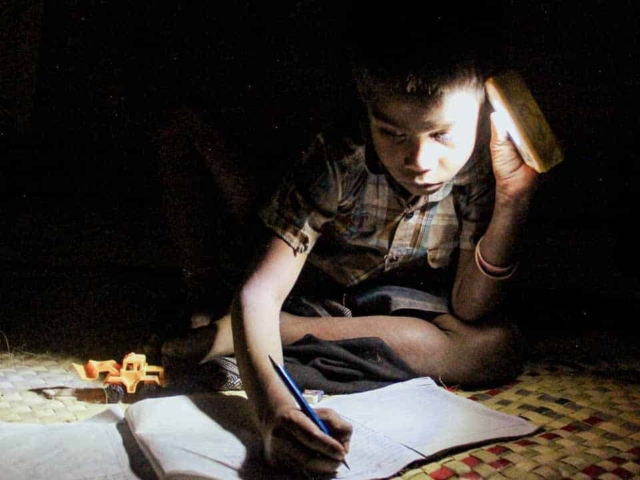 Children in East Sumba studying by the light of a SolarBuddy lamp, seated on the floor of their wooden home, deeply focused on their homework.