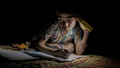 Children in East Sumba studying by the light of a SolarBuddy lamp, seated on the floor of their wooden home, deeply focused on their homework.