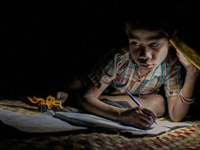 Children in East Sumba studying by the light of a SolarBuddy lamp, seated on the floor of their wooden home, deeply focused on their homework.