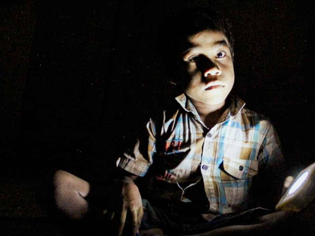 Children in East Sumba studying by the light of a SolarBuddy lamp, seated on the floor of their wooden home, deeply focused on their homework.