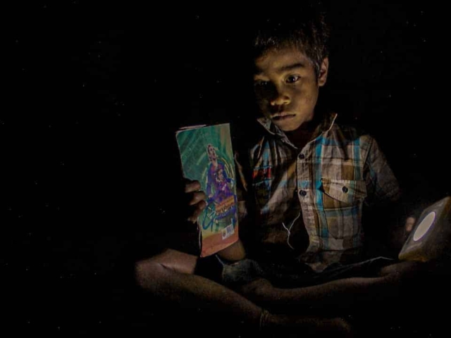 Children in East Sumba studying by the light of a SolarBuddy lamp, seated on the floor of their wooden home, deeply focused on their homework.