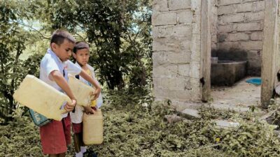 Children in rural Indonesia carrying water containers through dry, dusty paths to their home
