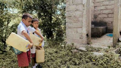 Children in rural Indonesia carrying water containers through dry, dusty paths to their home
