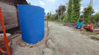 A blue water tank beside a village home with two residents seated nearby