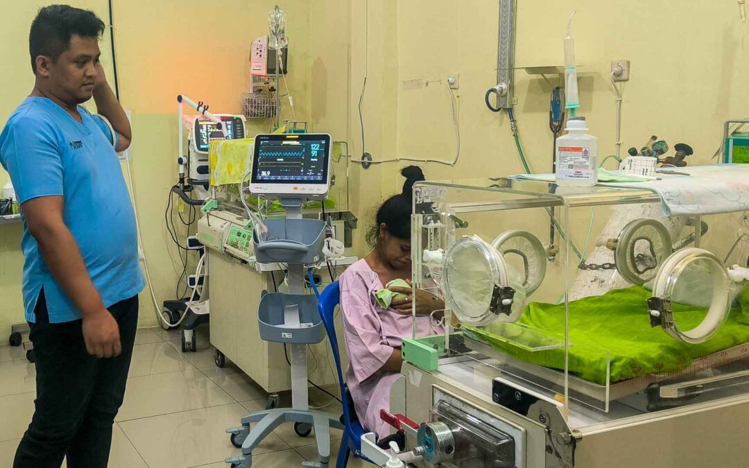 Neonatal unit in East Sumba’s only hospital: a mother with her newborn, a nurse monitoring equipment, and empty incubators, showing the critical lack of basic medical supplies.