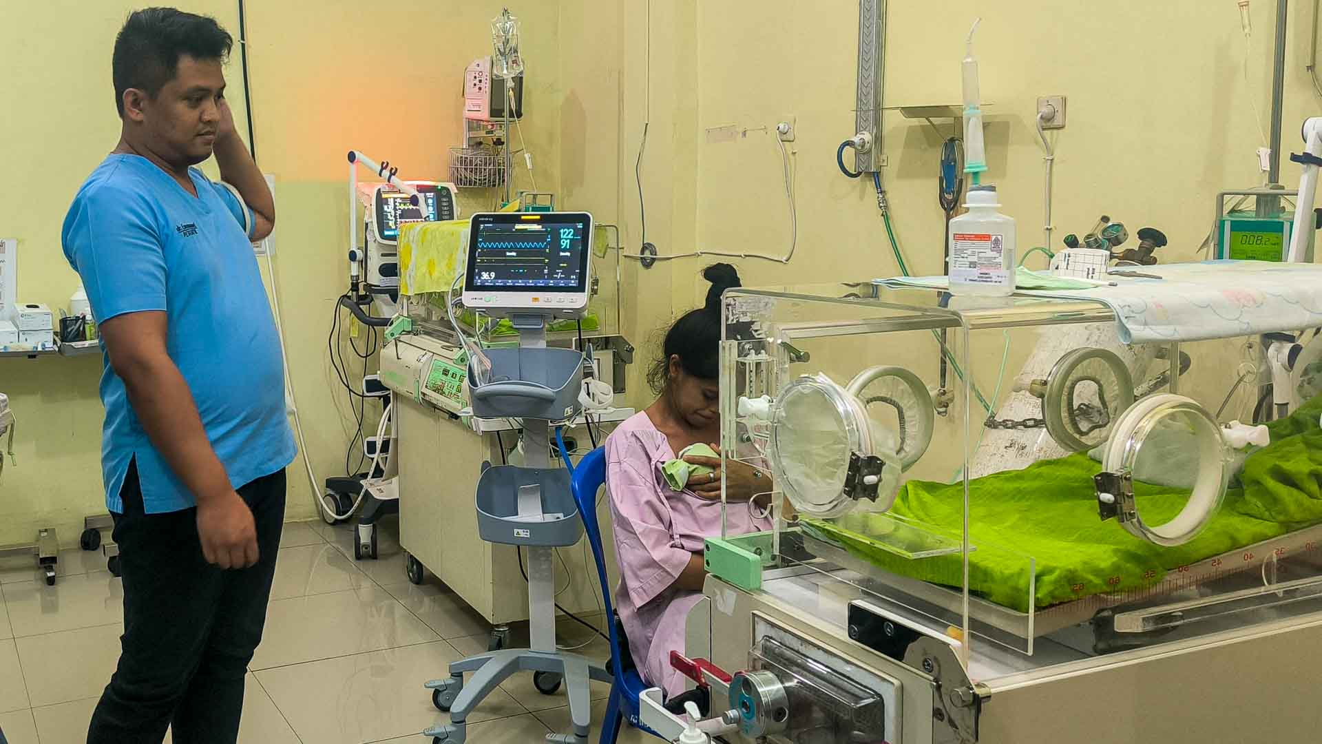 Neonatal unit in East Sumba’s only hospital: a mother with her newborn, a nurse monitoring equipment, and empty incubators, showing the critical lack of basic medical supplies.