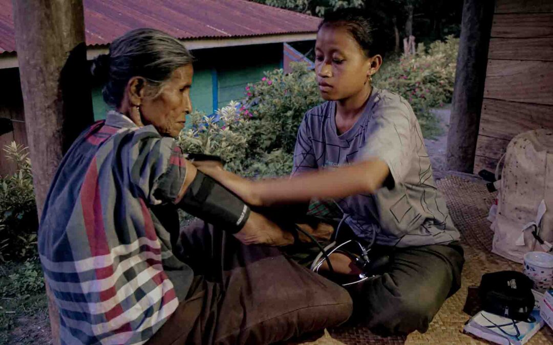 A Kawan Sehat health agent takes the blood pressure of an elderly woman in a rural village, sitting on a woven mat outside a wooden house at dusk, using a manual sphygmomanometer.