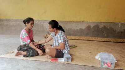 Katrina, Health Agent in a Village Without Roads • The Fair Future Foundation Katrina, a Kawan Sehat health agent, checking a woman’s blood pressure in a remote village in Lapinu, East Sumba, sitting on a bamboo mat with her medical kit beside her.