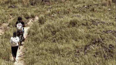Deux agentes de santé Kawan Sehat, l’une à pied, l’autre à moto avec un sac médical, suivent un étroit sentier au milieu des collines herbeuses vers un village isolé.
