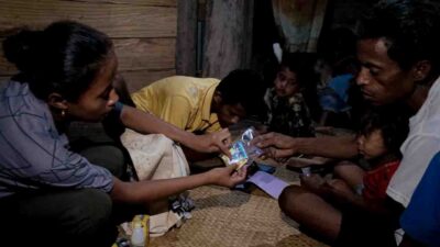 Kawan Sehat health worker Sarlota sits on the floor in her house in Lahiru, explaining a packet of medicines to a father while children watch beside them.