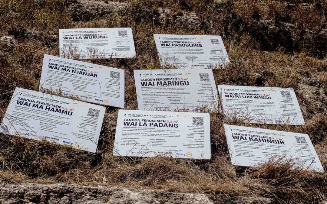 Nine laminated nameplates of water tanks are placed on dry grass in Hambarita, East Sumba. Each sign shows the name, capacity, coordinates, and donors of ferrocement reservoirs built by Fair Future Foundation and partners.