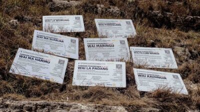 Nine laminated nameplates of water tanks are placed on dry grass in Hambarita, East Sumba. Each sign shows the name, capacity, coordinates, and donors of ferrocement reservoirs built by Fair Future Foundation and partners.