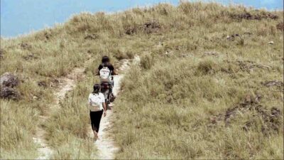 Two Kawan Sehat health agents on a dirt path in the Sumba savannah, one walking barefoot and one on a motorbike, on their way to provide medical care in a remote village.