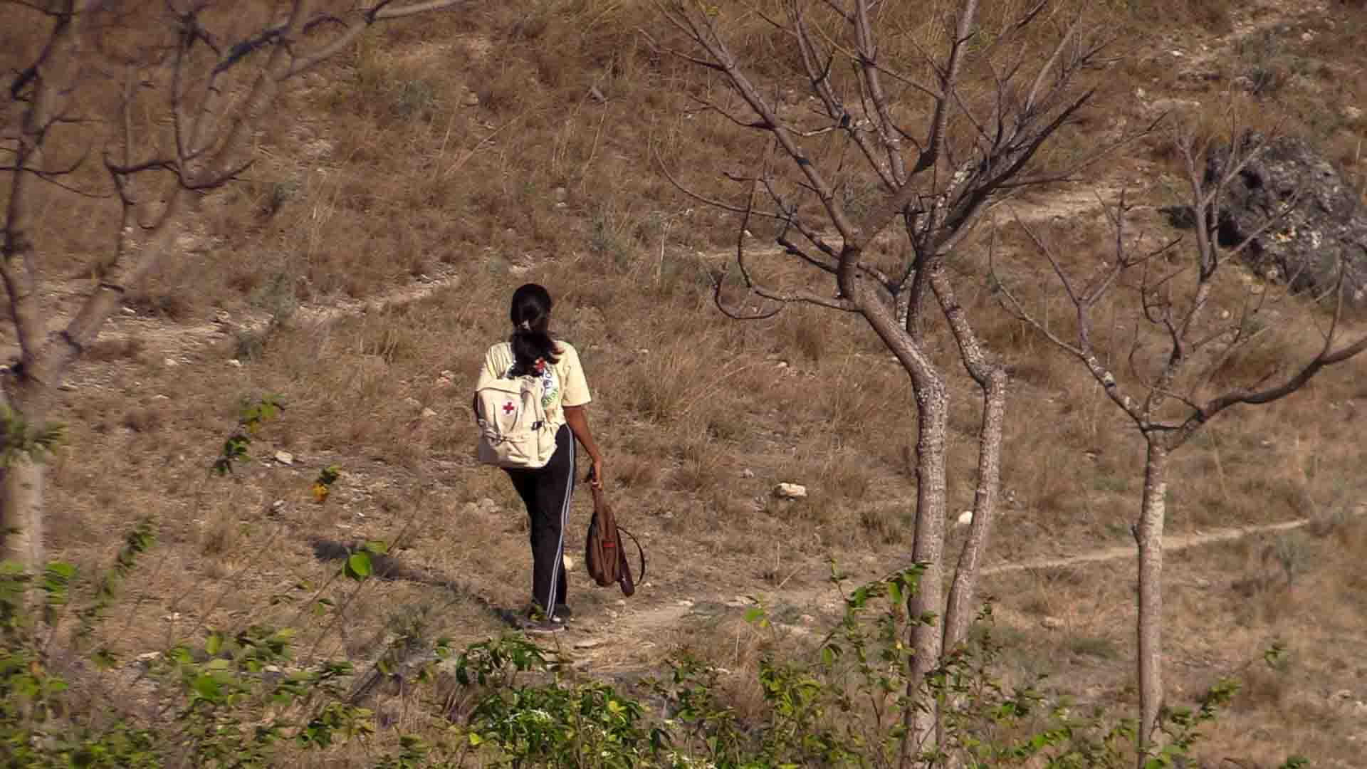 Kawan Sehat health agent walking through dry hills of East Sumba with a medical backpack to deliver care to remote villages.