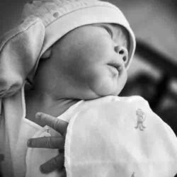 Black and white close-up portrait of newborn Sorai looking to the side, wrapped in a blanket, with an adult hand gently holding him.