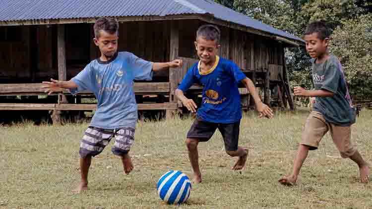 Trois garçons jouent au football pieds nus devant une maison en bois dans un village isolé de l'est de Sumba, en Indonésie.