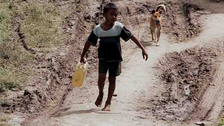 Un jeune enfant transporte un récipient d'eau le long d'une route rurale aride dans l'est de Sumba, en Indonésie, illustrant le défi quotidien que représente l'accès à l'eau potable dans les villages reculés.