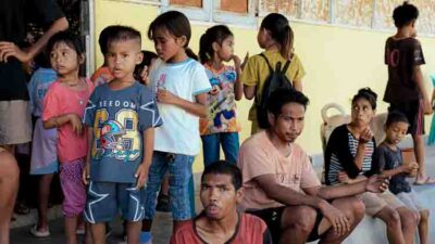 Group of children and a few adults waiting in line along a yellow wall, some seated and chatting.