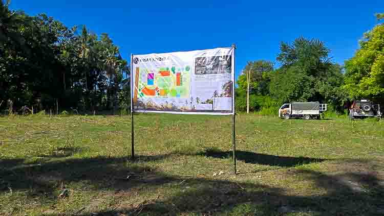 Swiss non-profit medical organisation working in ultra-rural indonesia Informational sign on two posts in an open grassy field, with trees and a bright blue sky behind; a few trucks are parked in the background.