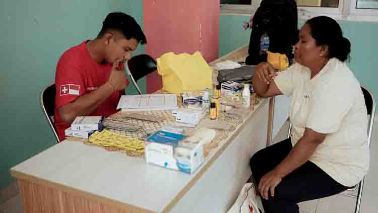 Swiss non-profit medical organisation working in ultra-rural indonesia Healthcare worker in a red shirt writes on forms at a clinic desk; patient sits opposite amid boxes and medical supplies.
