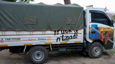 Side view of a white cargo truck with a gray tarp, a colorful mural on the cab, and banners/logos on the side (including 'Truck n Load!'). The vehicle is parked on a dirt surface.