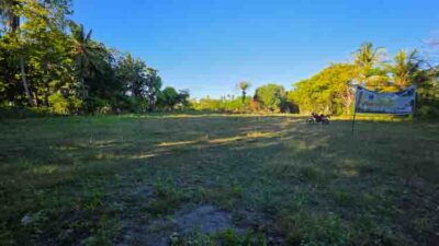 Open grassy field with trees along the left edge, a banner on the right, and a motorcycle parked in the distance under a clear blue sky.