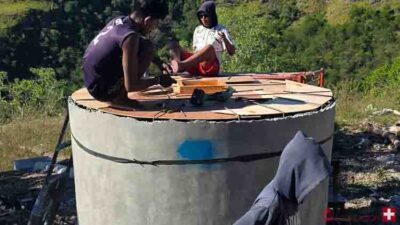Two people work on a large circular concrete water tank lid outdoors, using wooden boards and tools in a grassy, wooded area.