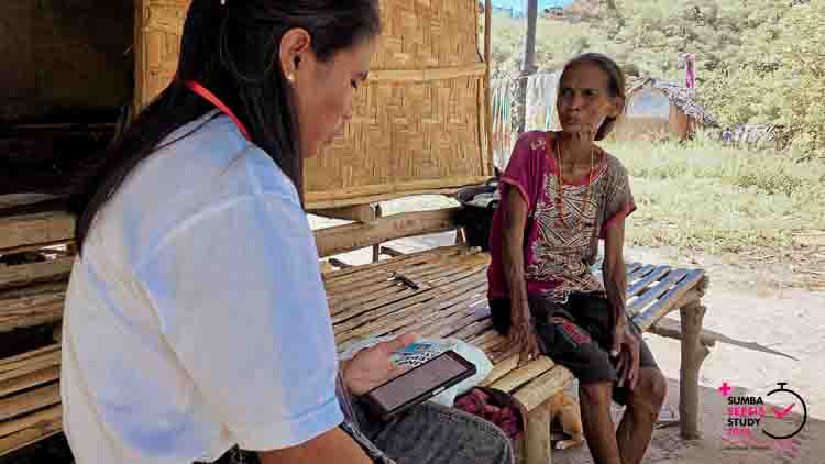 A field investigator interviews a woman in a remote village in East Sumba for the Sumba Sepsis Study 2026, using a tablet to collect medical data on infections and delays in care.