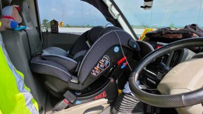 Infant/child car seat installed in the front passenger seat of a vehicle, with steering wheel and dashboard visible.