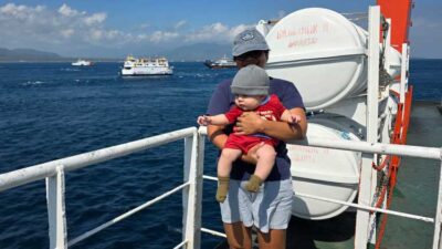 Adult holding a baby on a boat deck, with a large life raft behind and the ocean in the background.