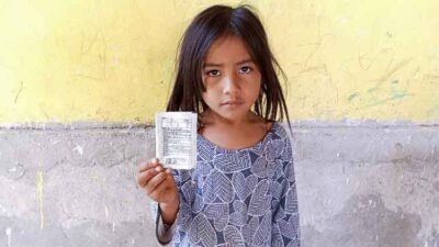 Young girl in a patterned blue dress holds up a small packet toward the camera, with a somber expression against a yellow wall.