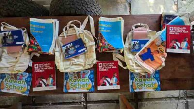 Row of beige backpacks hung on a wooden rail with health brochures and 'Kawan Sehat' posters nearby.