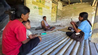 Family gathered in a simple bamboo hut; mother with baby on the left, man sitting in the back, and woman on the right using a smartphone.