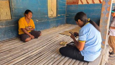 Two women sit on a wooden veranda; one wears a yellow shirt, the other in blue, weaving a mat or basket.