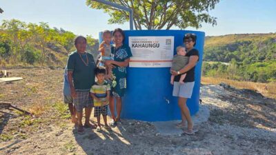 People stand beside a large blue water tank with a label reading KAHUNGU; two women hold babies, with several children nearby, in a dry outdoor setting.