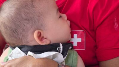 Sleeping infant resting on a caregiver's shoulder, with a dark bandana, in a red shirt with a white cross visible beside them.