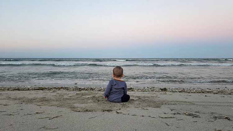 Seven-month-old Sorai sitting on the sand with his back to the camera, looking across the waves and horizon in East Sumba.