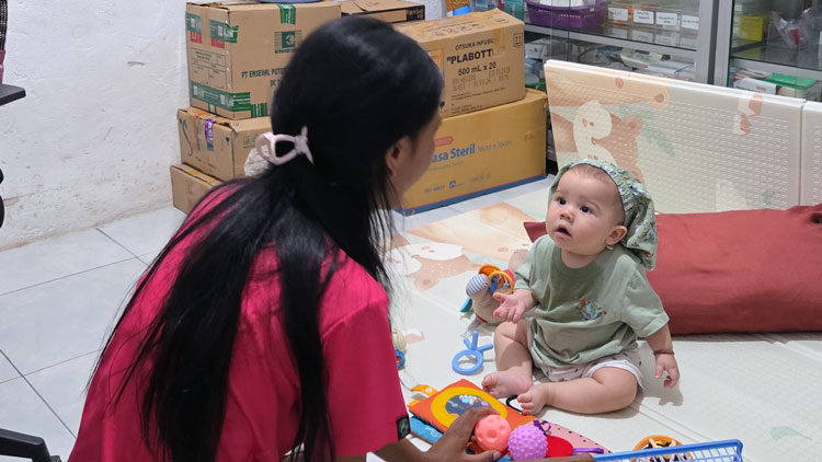 Sorai sitting on a play mat with Susanti inside the pharmacy of Rumah Kambera 1.0, surrounded by boxes of medicines and medical supplies in East Sumba.